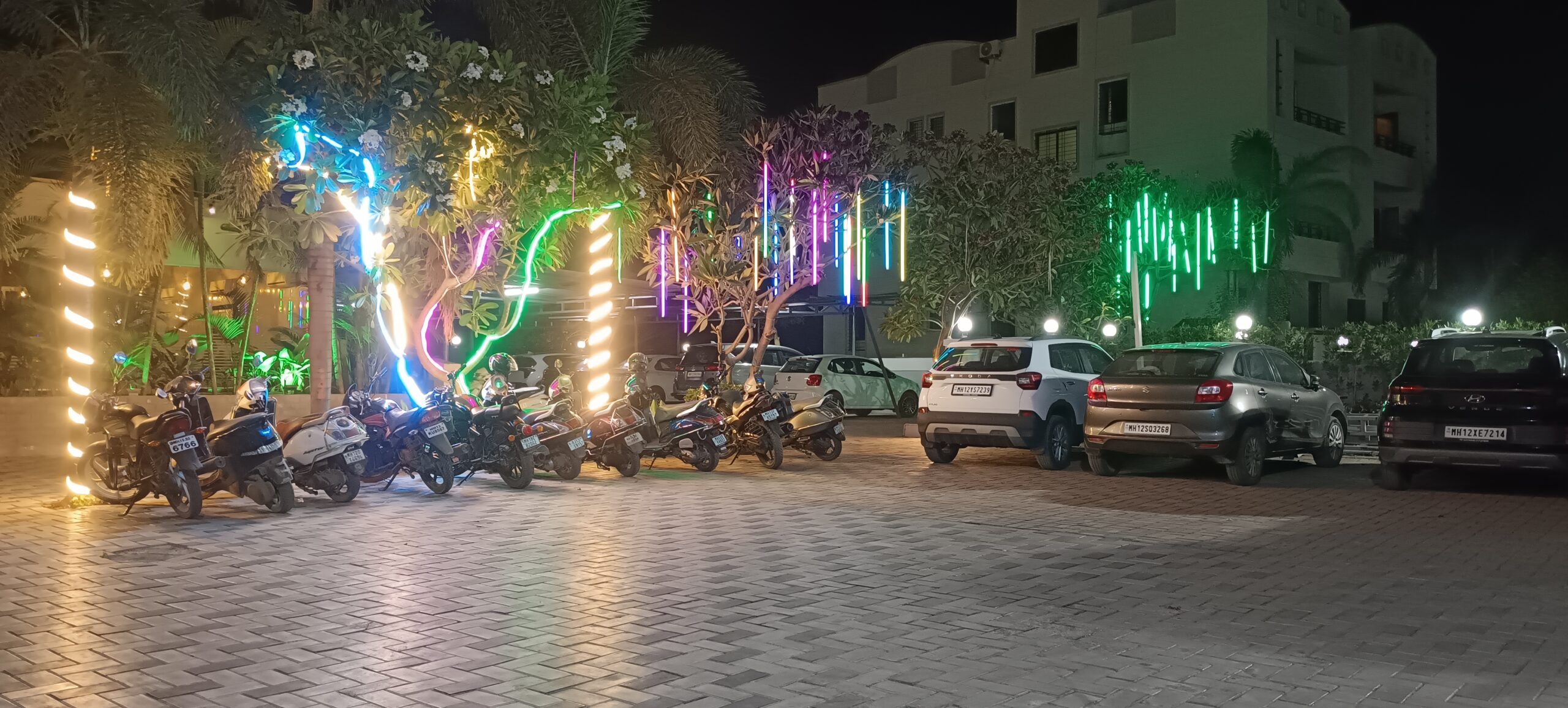 Colorful neon decorative lights on trees at Shivrudra Hotel parking area with parked bikes and cars at night Party area, venue, halls